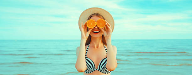 Summer portrait of happy smiling woman holding oranges covering her eyes on the beach on sea background stock photo