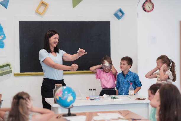 istock Elementary School Science Classroom: Enthusiastic Teacher Explains Chemistry to Diverse Group of Children, Little Boy Mixes Chemicals in Beakers. Children Learn with Interest 1412269548