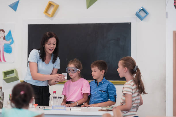 istock Elementary School Science Classroom: Enthusiastic Teacher Explains Chemistry to Diverse Group of Children, Little Boy Mixes Chemicals in Beakers. Children Learn with Interest 1412269704