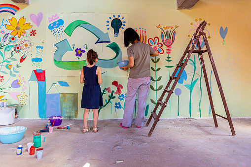 Creative girl with her mother painting with acrylic paints mural in the wall