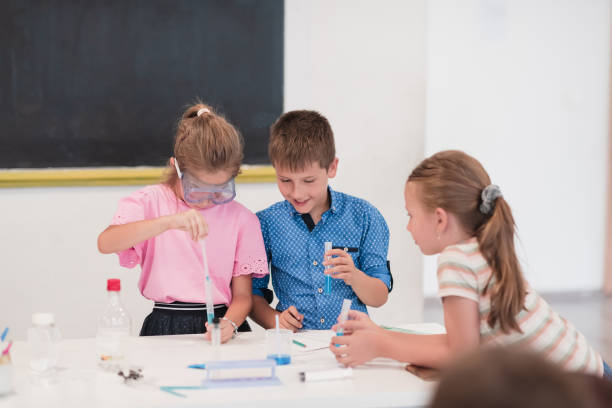 istock Elementary School Science Classroom: Enthusiastic Teacher Explains Chemistry to Diverse Group of Children, Little Boy Mixes Chemicals in Beakers. Children Learn with Interest 1412270649