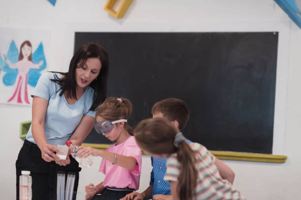 istock Elementary School Science Classroom: Enthusiastic Teacher Explains Chemistry to Diverse Group of Children, Little Boy Mixes Chemicals in Beakers. Children Learn with Interest 1412271370