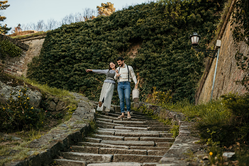 Spontaneous and loving photo of a beautiful young adult couple enjoying their weekend out, in a fortress, walking down the old stone stairs with a joyful smiles. A beautiful brunet is being playful, dancing with a wind while enjoying conversation, and holding hands with her handsome bearded partner that's carrying a cute white wooden basket, prepared for a picnic. Both retro stylishly dressed.
