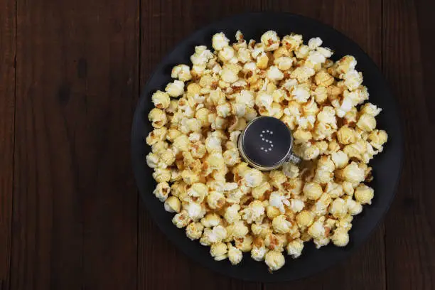 Overhead shot of a salt shaker in a shallow bowl surrounded by fresh made popcorn. Horizontal format on rustic dark table with copy space.