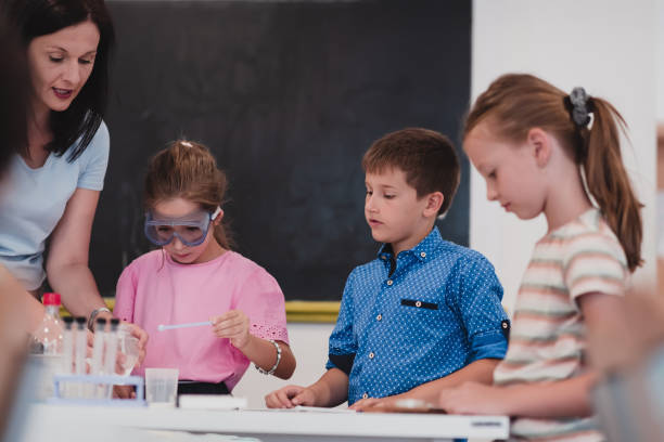 istock Elementary School Science Classroom: Enthusiastic Teacher Explains Chemistry to Diverse Group of Children, Little Boy Mixes Chemicals in Beakers. Children Learn with Interest 1412274437