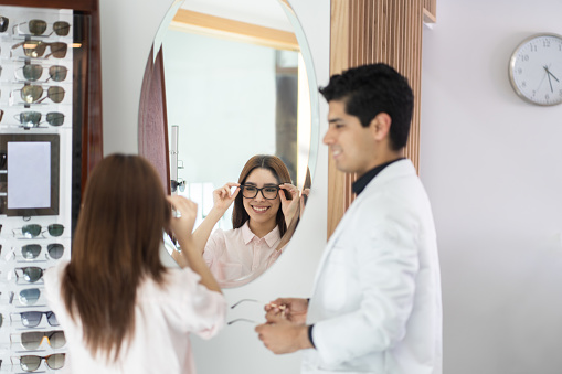 A latin cheerful young woman trying on eyeglasses with an ophthalmologist and looking herself in the mirror.