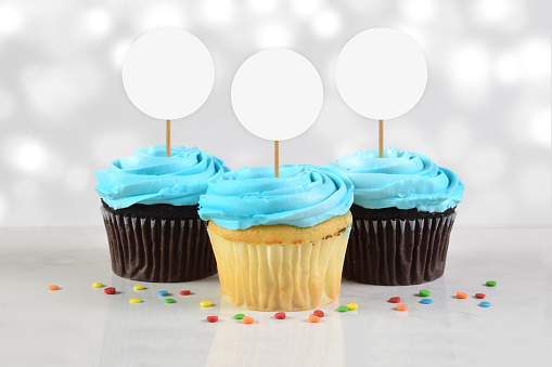 This cupcake topper mockup features three delicious blue frosted cupcakes atop a white marble background dusted with colorful sprinkles. The glowing white backround lights add a touch of class to this cupcake mock up.