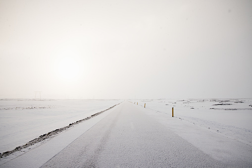 Snow covered icelandic Country Road in Winter against the setting sun close to dusk shining through a snow cloud wall sky. Frozen Icelandic Country Road - Highway hrough the Highlands close to Hekla Volcano in Winter. Iceland, Northern Europe, Nordic Countries.