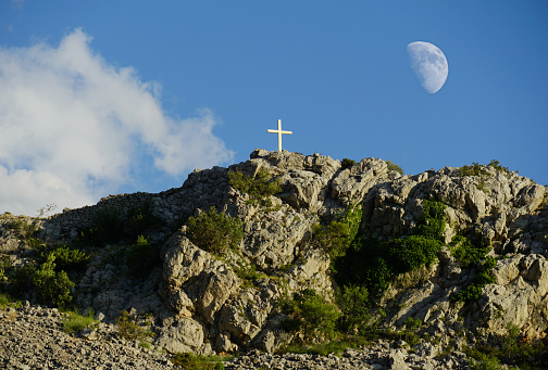 A white cross on top of a huge rock under the blue sky and the moon in the sky