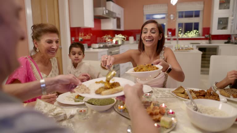 An over-the-shoulder view of a family get-together in their home. They are having traditional food for Diwali celebrations and passing food to each other at the table.
