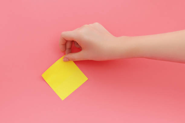 Woman's hand glues yellow sticker or sticky notes on pink background.