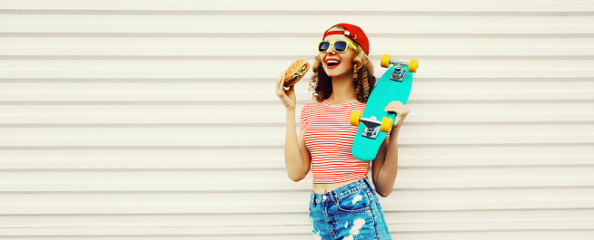 Portrait of stylish young woman with burger and skateboard wearing baseball cap, sunglasses on white background