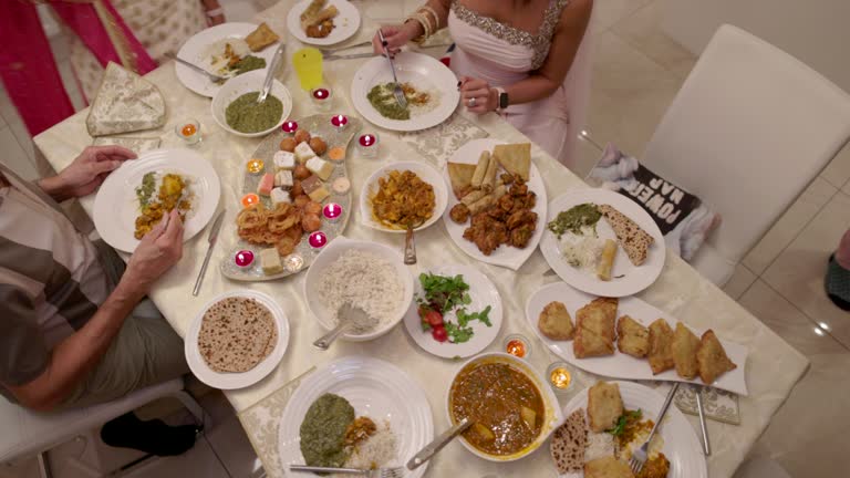 An aerial panning view of a family all sitting together to sit down for some freshly cooked food in their living room as they celebrate Diwali together