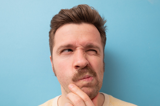 Young man with funny mustache having doubts and with confuse face expression on isolated blue background