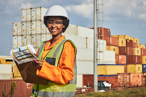 A young African woman fore person working in a shipping container store yard and wearing personal protective wear and using a clipboard and assessing containers