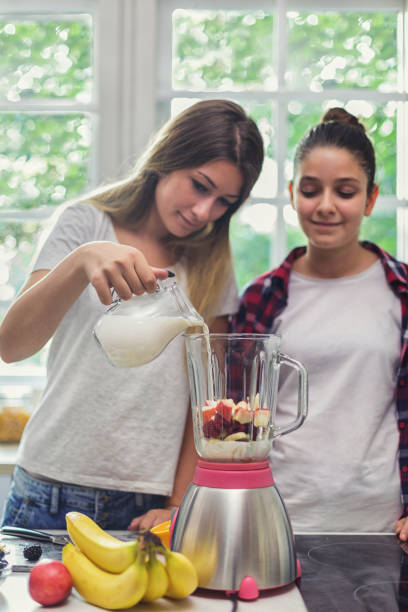 two happy young girls making fresh organic smoothie using blender in the kitchen - blender apple banana color image imagens e fotografias de stock