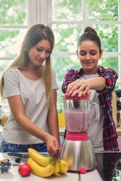 two happy young girls making fresh organic smoothie using blender in the kitchen - blender apple banana color image imagens e fotografias de stock