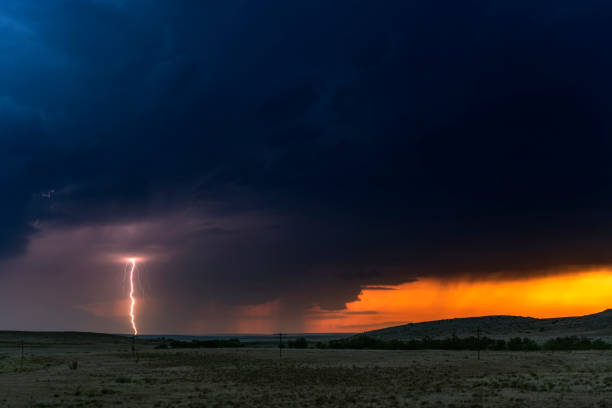 A large lightning strike at dusk in an open plain framed against a deep, dark orange sunset and stormy skies.