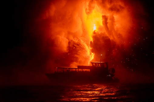 A tourist boat floats in front of exploding lava at night, giving people a view of mother nature in her fury.