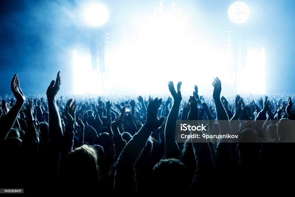 Performance of a popular group. The crowd with raised hands against the stage light. Performance of a popular group. The crowd with raised hands against the stage light Nightlife Stock Photo