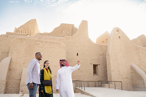 Front view of Saudi guide in traditional attire and mid adult couple standing together in front of Salwa Palace as they explore open-air museum. Property release attached.