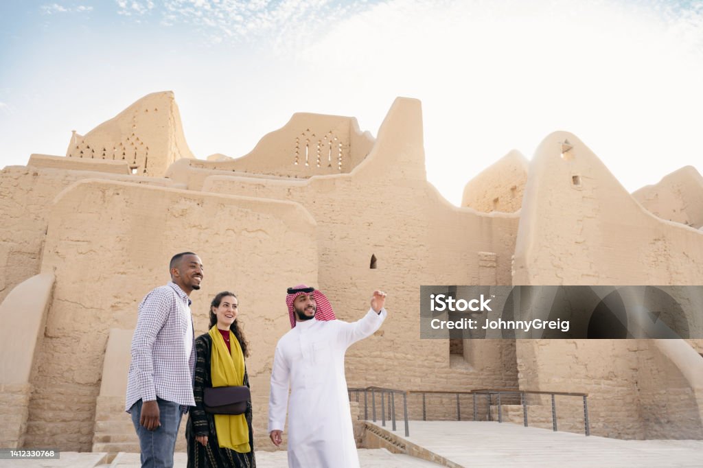 Tourists admiring restored ruins of At-Turaif near Riyadh Front view of Saudi guide in traditional attire and mid adult couple standing together in front of Salwa Palace as they explore open-air museum. Property release attached. Saudi Arabia Stock Photo