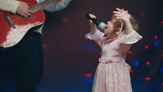 Little girl on stage in vintage dress, she sings into microphone and dances, her father plays acoustic guitar. Color music is shining and smoke is billowing. Father and Child Day, performance on stage
