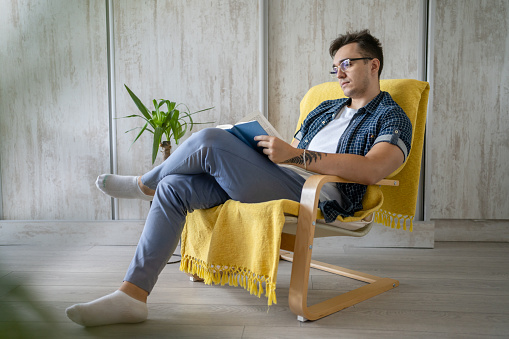 One man caucasian male young adult sitting in a chair at home reading a book wear blue shirt and wear eyeglasses leisure time concept education copy space