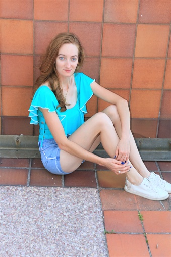 A model from Paraguay sitting on a brick floor with her knees up. She is wearing a blue sleeveless top, denim shorts and white shoes. She also has a necklace and a ring on.