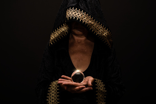 Witch in black dress with face covered by hood holding crystal ball in hands. Space for text. Halloween or Day of the Dead concept