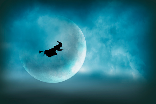 A witch flying on her broom across the sky is silhouetted in front of a large moon on Halloween night.