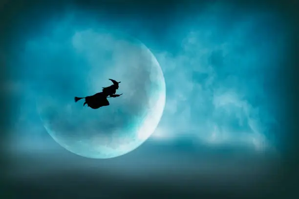 A witch flying on her broom across the sky is silhouetted in front of a large moon on Halloween night.