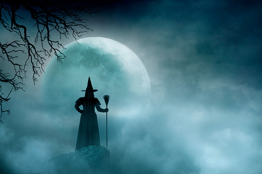 A rear view of a witch holding her broom stick as she stands on a rocky promontory and looks out toward the moon shrouded in clouds in the distance.