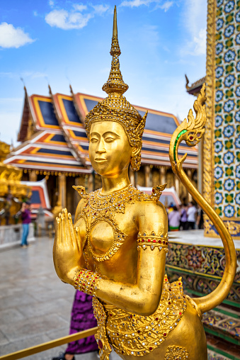 View of Buddha sculpture Kinora or Kinnaree ( mythological creature, half bird, half man ) at Wat Phra Kaeo and Grand Palace in Bangkok, Thailand. Many details of Grand Palace in the background, also visible are beautiful cloudscape with blue sky and cumulus clouds on one sunny day in Bangkok.