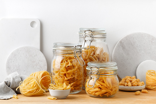 A variety of pasta in glass jars on a wooden table. White wall. Assortment raw pasta in a glass jars. Food set. Modern wooden kitchen table background. Scandinavian style. Mock up.