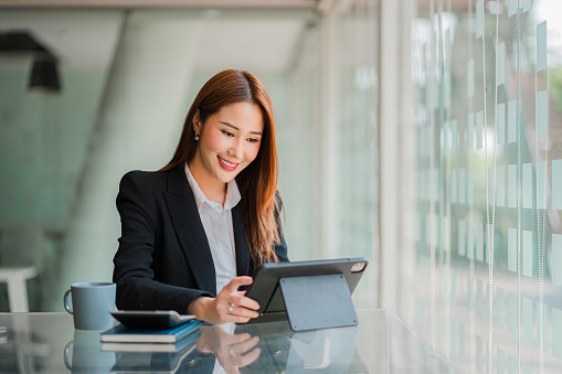 Beautiful smiling Asian businesswoman working using a tablet at the office while sitting in a modern office successful business idea