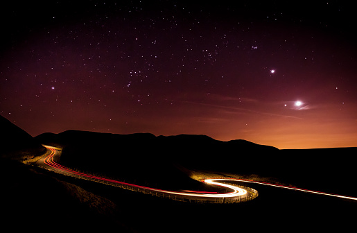 Light trails and stars cape with Venus, Jupiter, Orion and the moon clearly visible above a winding road in the Peak District National Park. Derbyshire, England, United Kingdom, Europe
