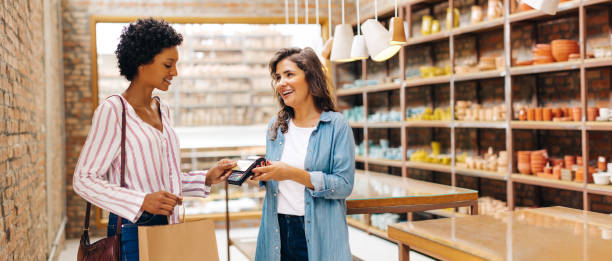 Happy shop owner receiving a contactless credit card payment from a customer Happy ceramic store owner receiving a contactless credit card payment from a customer in her shop. Successful small business owner smiling cheerfully while serving a customer. b2b sales stock pictures, royalty-free photos & images