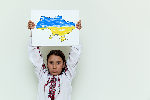 A girl in an embroidered dress stands against the background of a white wall, holding a white sheet with a picture and an inscription. The girl protests against the war.