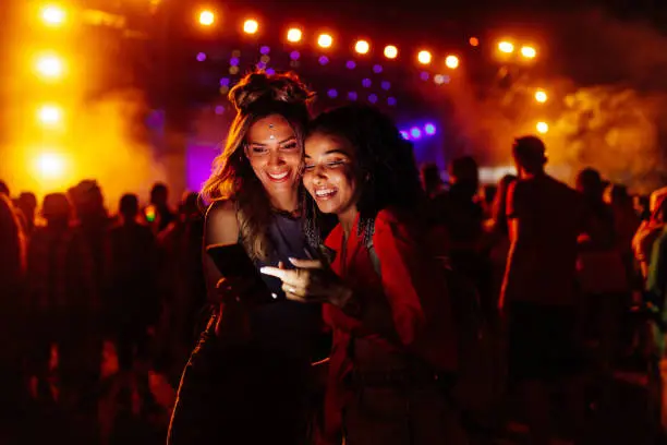 Two biracial women in a crowd at a concert using cellphone together