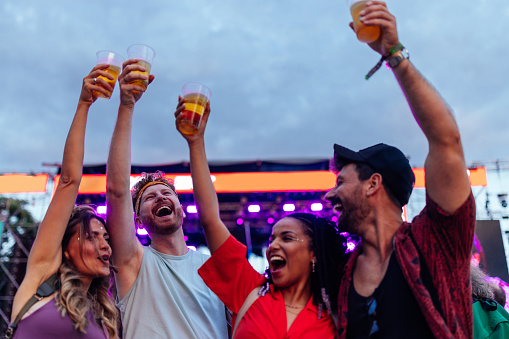 Excited four multiracial friends enjoying a concert performance of a unrecognizable artist. Raised hands of fans with glasses of beer are in the focus against purple lit stage.