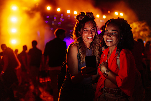 Biracial female friends standing in the crowd at music festival and taking selfie via cellphone