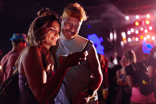Adorable young couple standing in crowd at music festival, looking at cellphone, searching for line up