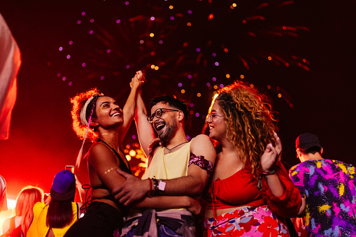Young multiracial friends dancing at music festival. They are jumping with their arms raised illuminated with red stage light and with fireworks in the background