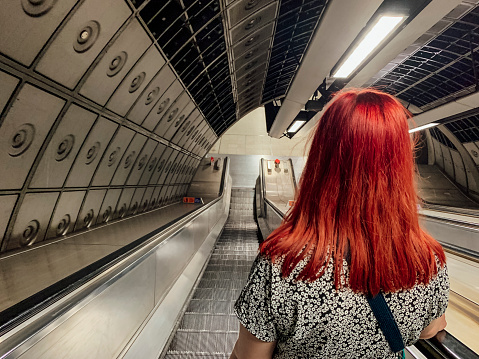 A rear view, medium shot of an unrecognisable female university student with dyed red hair travelling down an escalator on the London Underground.