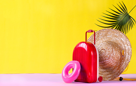 Background with red suitcase, rubber ring and straw hat on yellow. Travel composition. Summer vacation concept. Mockup with copy space