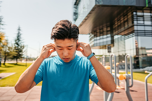 A handsome young Chinese athlete man putting on wireless headphones to listen to music while working out in a park outdoors.