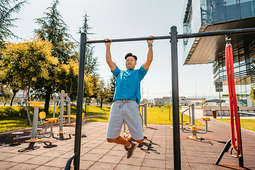 A handsome young Chinese man doing pull-ups on a bar outdoors in a public park.