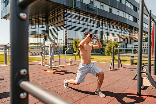 A handsome young Chinese man training outdoors by doing lunges in a public park. Wearing a VR headset.