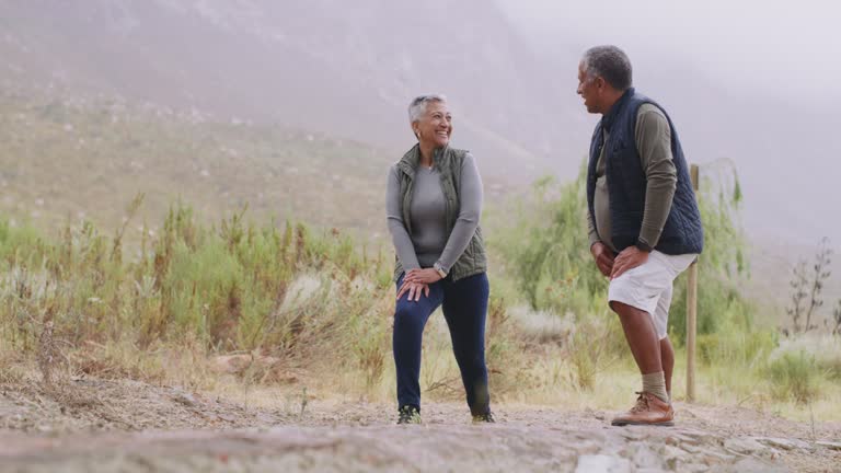 Mature mixed race couple stretching their legs before a mountain hike. Retired senior couple bonding, talking before hiking in nature. Fit, active older couple getting to hike together